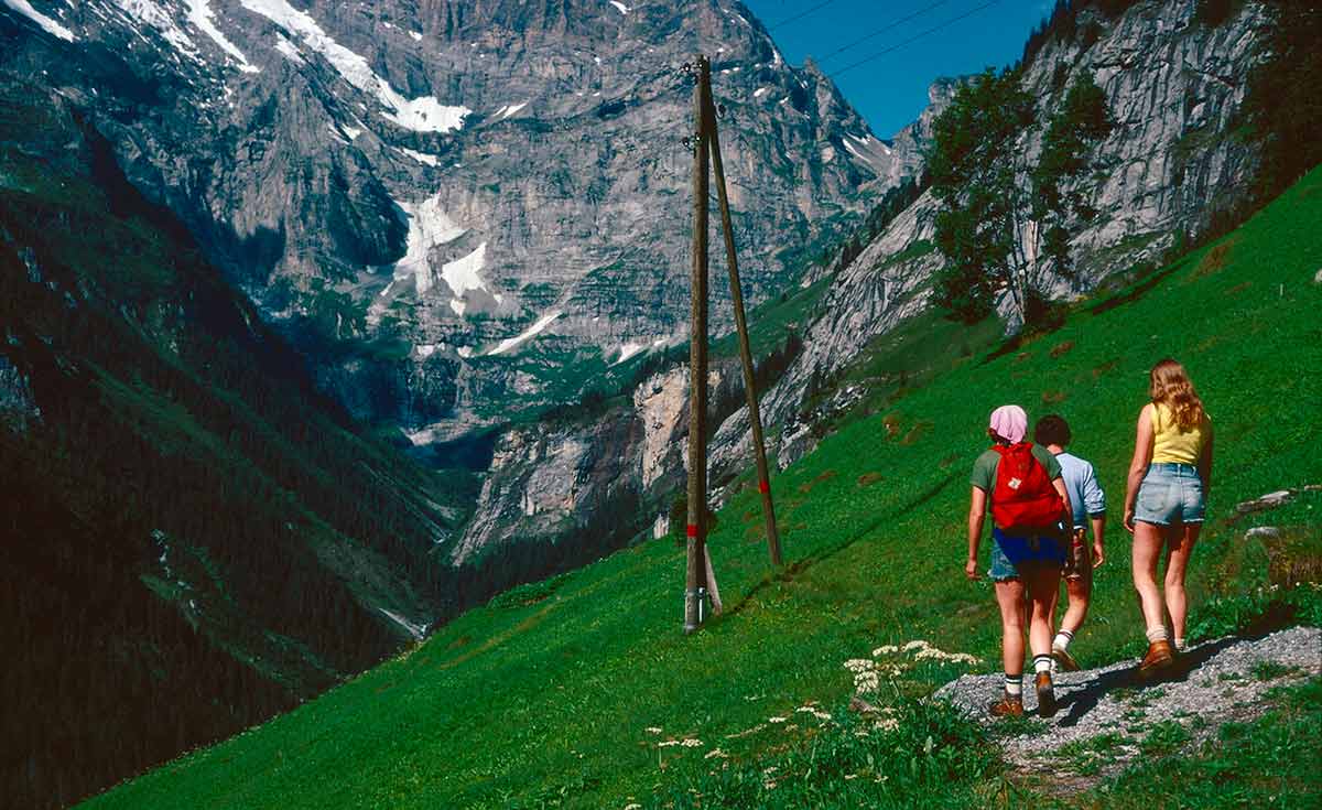 People walking across a steeply sloping meadow, mountains rise in the background