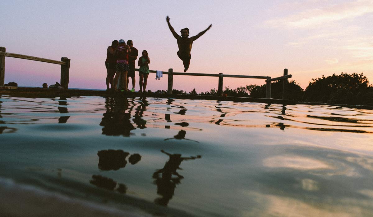 Several people standing by a pool at dusk, one person jumping into the water