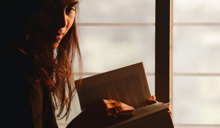 Woman sitting in window, reading a book and looking towards camera