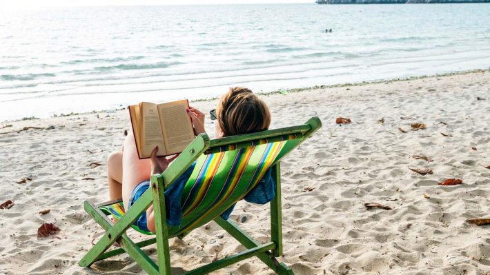 Woman reading a book, sitting in a folding chair on a beach by the sea