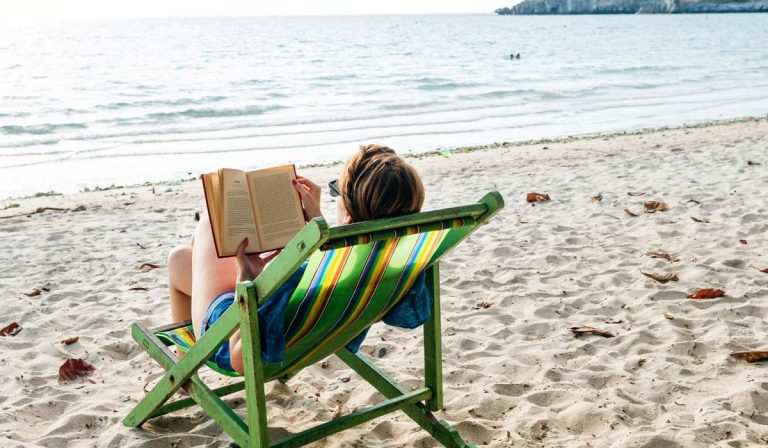 Woman reading a book, sitting in a folding chair on a beach by the sea